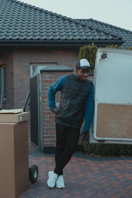 A man dressed in a black padded vest, blue long-sleeve shirt, and black trousers, wearing a white cap and white shoes, is standing outdoors next to a white moving van with a wooden panel on the side. He is leaning slightly against the open rear door of the van, which is parked on a paved driveway in front of a residential house with a tiled roof and brick walls. The man has one hand on his hip and appears to be resting during a home relocation process. Nearby on the driveway, there is a large cardboard box on a dolly wheel, indicating packing and moving activities. The scene is illuminated by natural daylight, and the surrounding environment includes a small bush and part of the house, suggesting a typical suburban setting. This image is associated with removals and furniture transport services provided by Removal Companies Harringay, emphasizing loading, packing, and home relocation logistics.