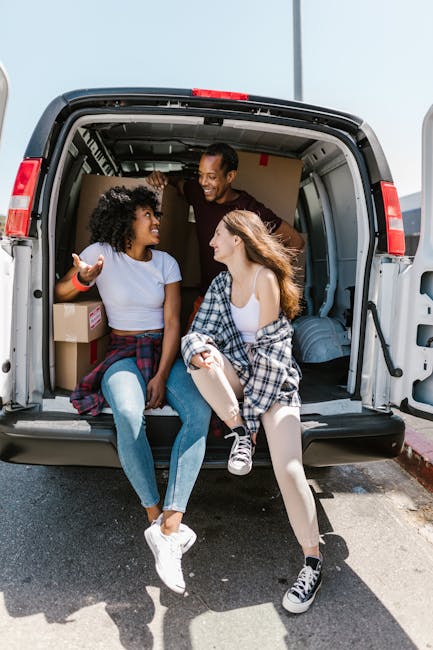 Two young women are sitting on the edge of an open moving van, engaging in conversation and smiling. One woman has curly hair, is wearing a white t-shirt and jeans, and has a plaid shirt tied around her waist. The other woman has straight hair, is dressed in a white top and patterned pants, and has a plaid shirt draped over her shoulders. Behind them, inside the van, visible packing boxes made of cardboard with labels are arranged alongside rolled-up blankets or cushions. The van’s interior shows some metal rails and a roof lining, indicating it is being used for a home relocation or furniture transport. The scene is outdoors on a paved street, with natural daylight illuminating the area, suggesting a moving day in progress. The image captures a moment of packing and loading activities as part of a professional removals service, exemplified by the open back of the van and the casual, friendly interaction between the two individuals.