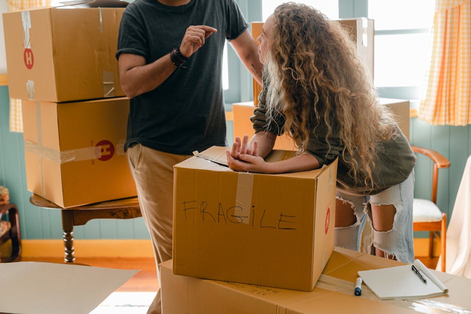 A man and a woman are inside a room during a home relocation process, surrounded by cardboard boxes, some sealed with packing tape and marked with a red 'H' symbol. The man, with dark hair and wearing