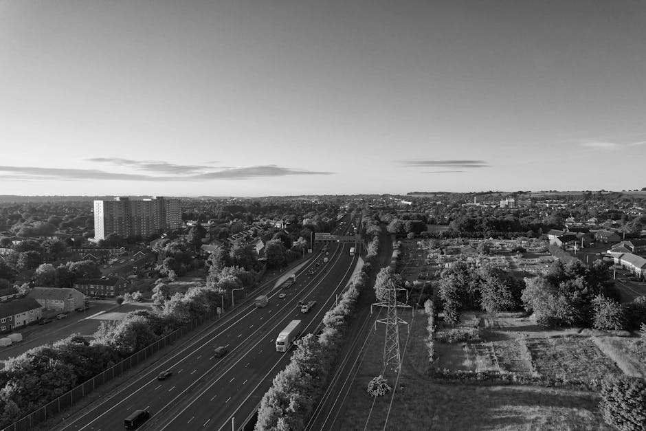 A black-and-white aerial photograph showing a multi-lane road with moving vehicles, including cars and a large van, situated next to residential houses and green spaces. The road is bordered by a fence on one side and power lines with tall pylons running parallel along the outskirts of the urban area. In the background, there are low-rise apartment blocks, scattered trees, and open land extending to the horizon. The scene captures a typical urban and suburban landscape during daylight hours, illustrating the type of environment where home relocation and furniture transport services provided by Removal Companies Harringay might operate, highlighting the logistical context of packing, loading, and street-level moving activities involved in house removals.