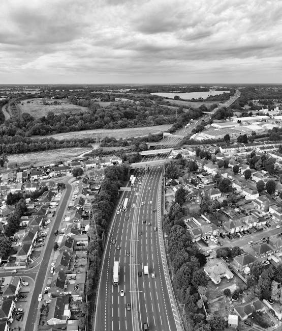A black-and-white aerial photograph showing a busy urban and suburban area with a multi-lane motorway running through the centre. The motorway is filled with various vehicles, including cars and trucks, and is flanked by residential houses, small commercial buildings, and patches of greenery. On the left side, there are closely packed houses with driveways and gardens, while on the right, more sparse residential properties and wider roads are visible. In the background, open fields, wooded areas, and a large body of water can be seen under a cloudy sky. The image captures the scale of urban infrastructure and the surrounding landscape, which may be relevant for logistics and transportation considerations in house removals. This scene is relevant to services like those provided by Removal Companies Harringay, who facilitate the packing and moving process related to home relocation and furniture transport.