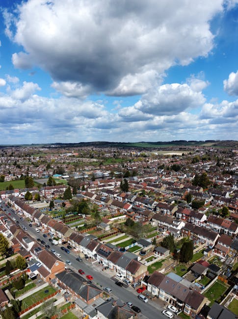 A black-and-white aerial photograph showing a multi-lane road with moving vehicles, including cars and a large van, situated next to residential houses and green spaces. The road is bordered by a fence on one side and power lines with tall pylons running parallel along the outskirts of the urban area. In the background, there are low-rise apartment blocks, scattered trees, and open land extending to the horizon. The scene captures a typical urban and suburban landscape during daylight hours, illustrating the type of environment where home relocation and furniture transport services provided by Removal Companies Harringay might operate, highlighting the logistical context of packing, loading, and street-level moving activities involved in house removals.