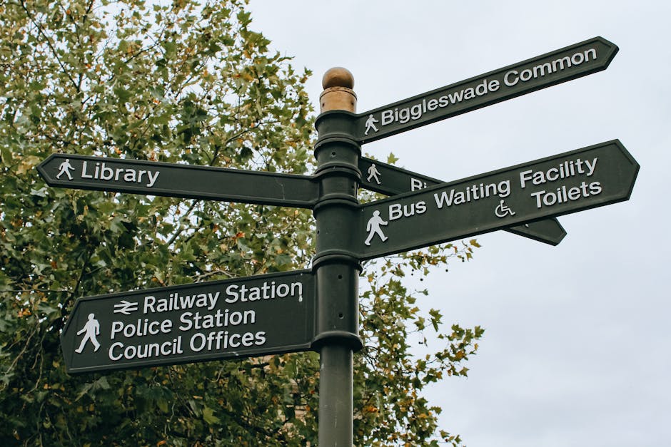 A black multi-directional street signpost featuring white engraved text and icons indicating various local destinations including Biggleswade Common, Library, Railway Station, Police Station, Council Offices, Bus Waiting Facility, and Toilets. The signpost is situated outdoors against a backdrop of leafy trees with green and brown foliage, under an overcast sky. The signs are mounted on a central pole with a rounded top, and each pointing in different directions to guide pedestrians. The image captures the signage in daylight, and the signpost is positioned on a street or public area where house removals and relocations might be centered. This scene is relevant for documenting local infrastructure in the context of home or furniture transport, as seen in professional removals services offered by companies like Removal Companies Harringay.