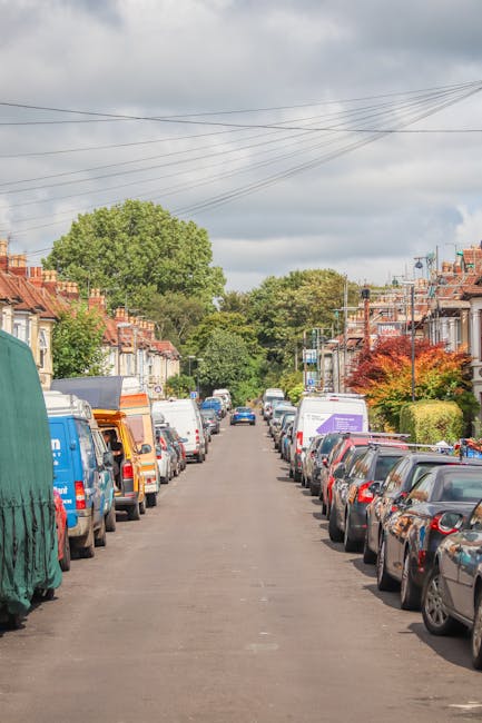 A residential street scene during daytime showing a row of parked cars on both sides of the narrow asphalt road, with some vehicles covered with protective tarps or plastic wraps. The street is lined