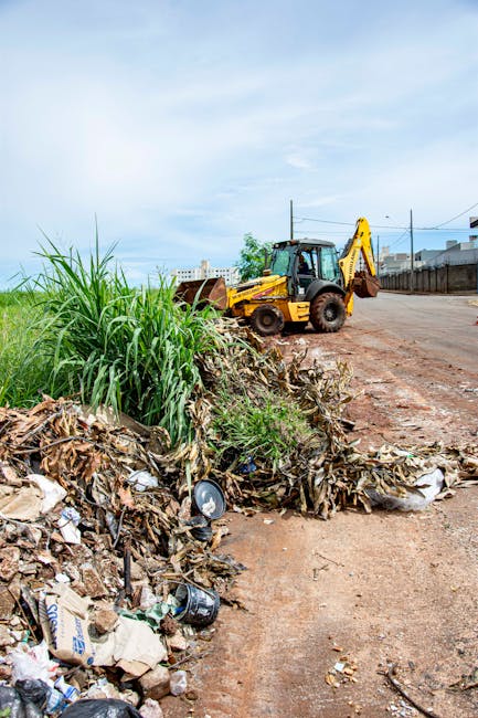 An outdoor scene showing a construction or cleanup area with a bright yellow backhoe loader positioned on a dirt road. In the foreground, there is a pile of mixed household waste and debris, including cardboard boxes, plastic bottles, and paper, along with some green foliage and overgrown grass. The debris appears to be temporarily stored on the side of the road, possibly for removal or disposal. The background features a clear blue sky with a few clouds, a wire fence on the right, and distant buildings or structures. The scene suggests ongoing site clearing or waste removal, which could relate to home or property renovation projects, in line with house removals or disposal services offered by Removal Companies Harringay.