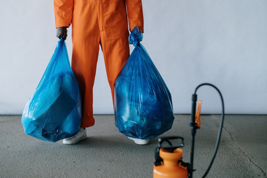 A person dressed in an orange work jumpsuit is standing inside a room with a plain, light-colored wall and a concrete floor, holding two large blue plastic garbage bags filled with household waste or items ready for disposal, with their hands gripping the bags by the tied tops. The person's face is not visible as the image focuses on their lower body and the bags. Nearby, a small portable sprayer with an orange tank and black handle is placed on the floor, suggesting preparation for cleaning or pest control during home relocation or furniture transport processes. The scene is well-lit, indicating it is daytime, and the environment appears to be part of a house or storage area where household items and waste are being sorted and organized for removals or disposal, consistent with services offered by Removal Companies Harringay related to house removals and waste disposal.