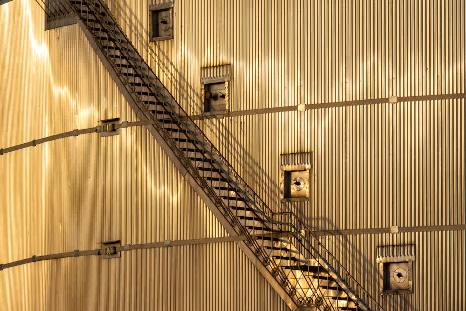 The image displays an external view of a building's metallic wall illuminated by warm sunlight, creating reflective highlights on its vertical panels. A metal fire escape staircase runs diagonally across the wall from the bottom left to the top right, with black steps and railings casting shadows on the surface. Several rectangular ventilation units are mounted on the wall near the staircase, and electrical conduit pipes run horizontally along the structure. The scene is lit naturally, emphasizing the industrial aesthetic. This setting showcases typical external features of urban buildings, relevant to home relocation and moving logistics, such as planning access routes for furniture transport, as detailed in the Harringay Ladder removals tips for narrow stair and access issues.
