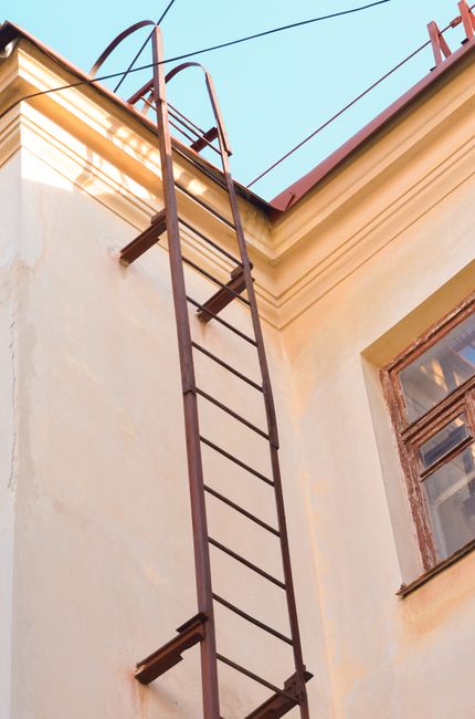 The image displays an external view of a building's metallic wall illuminated by warm sunlight, creating reflective highlights on its vertical panels. A metal fire escape staircase runs diagonally across the wall from the bottom left to the top right, with black steps and railings casting shadows on the surface. Several rectangular ventilation units are mounted on the wall near the staircase, and electrical conduit pipes run horizontally along the structure. The scene is lit naturally, emphasizing the industrial aesthetic. This setting showcases typical external features of urban buildings, relevant to home relocation and moving logistics, such as planning access routes for furniture transport, as detailed in the Harringay Ladder removals tips for narrow stair and access issues.