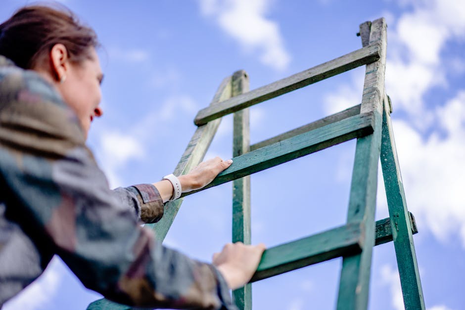 A person with brown hair, wearing a grey jacket and a white wristwatch, is seen from the side climbing a green wooden ladder outdoors under a partly cloudy sky. The ladder is positioned on the ground, leading upward, and the individual is holding onto the rungs with both hands. The scene is related to home relocation or moving processes, with the setting suggesting preparation or access for moving or packing activities, possibly in a residential area in Harringay. The image captures a moment during a packing and moving task, emphasizing the use of equipment like ladders in the logistics of furniture transport or house removals, consistent with professional services offered by companies such as Removal Companies Harringay.