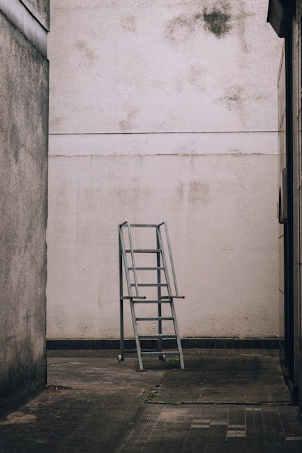 A silver metal ladder with four steps and safety rails at the top is positioned on a concrete outdoor surface, leaning against a plain, weathered white wall that has visible stains and discoloration. The setting appears to be an alleyway or side yard, enclosed by high walls on either side, with the wall behind the ladder being taller and featureless. The lighting is natural, suggesting daytime, with soft shadows cast by the ladder onto the ground. There are no other objects or furniture present in the scene. This image reflects a moment related to home relocation or moving preparation, possibly depicting the loading process or storage area in a house removal context, as may be relevant to services provided by Removal Companies Harringay, especially in relation to furniture transport and packing activities.