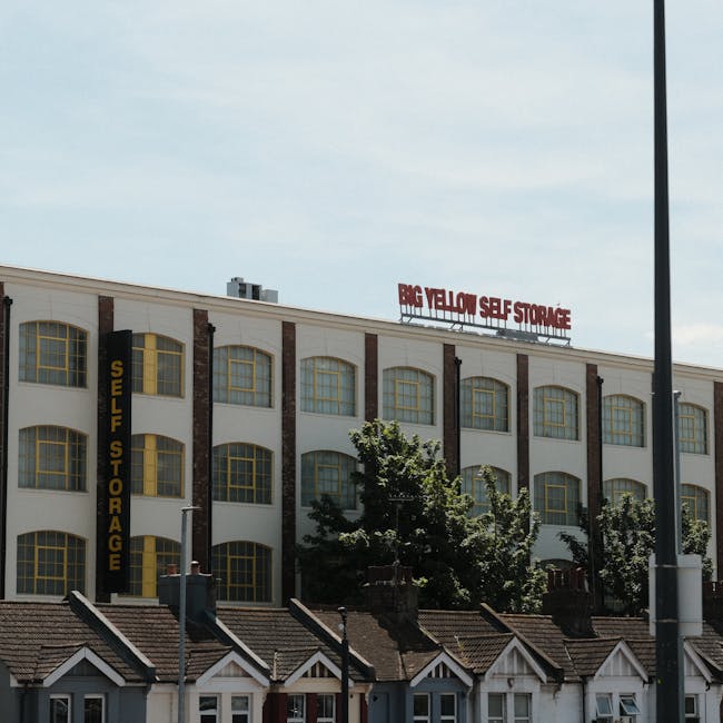 The image shows the exterior of a multi-story building with a sign on the roof reading 'Big Yellow Self Storage' in red letters, indicating a facility for storage services. The building features a white façade with brown vertical columns and large yellow-framed windows arranged in a grid pattern across all floors. On the side of the building, there is a vertical black sign with yellow text that reads 'Self Storage.' In front of the building, several small residential-style houses with pitched roofs and chimneys are visible, along with some trees and shrubs. A tall street lamp with a black pole stands to the right of the image. The overall scene depicts an urban environment during daylight with clear weather, suitable for a landscape showing commercial storage facilities involved in house removals, furniture transport, or packing and moving activities in the Harringay area. This visual context aligns with the services offered by Removal Companies Harringay for house relocation and storage solutions.