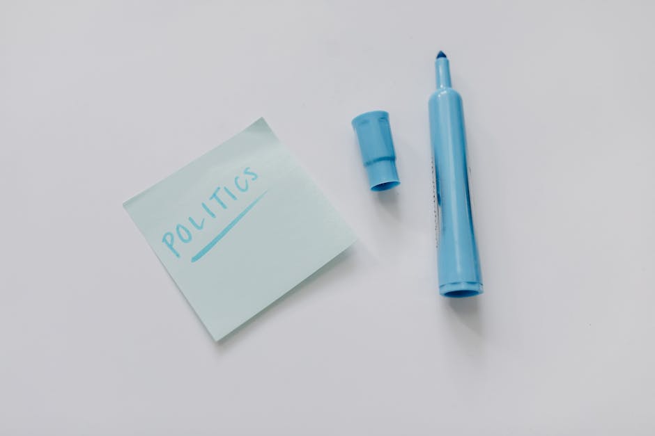A light blue sticky note with the word 'POLITICS' written in blue ink is placed on a clean, white surface. Next to the note, there is a matching light blue marker with the cap removed, revealing the tip, and the cap itself is positioned nearby. The marker is designed for writing on paper or sticky notes. The overall setting appears simple and uncluttered, suggesting a focus on note-taking or labeling activities. This image may be used to illustrate workspace organization or documentation in the context of house removals, packing, or moving logistics, emphasizing the importance of labeling and planning during a home or office relocation handled by Removal Companies Harringay.