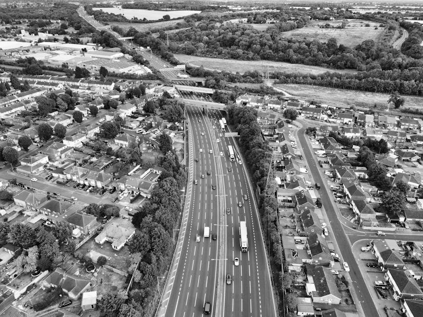 Aerial view of a suburban area with a major multi-lane road running through it, showing cars, trucks, and buses in motion. To the left of the road, there are numerous residential houses with back gardens, trees, and narrow streets. To the right, there is a row of terraced houses and parked vehicles along the roads. Beyond the roadway, the landscape includes open fields, patches of woodland, and distant farmland, with a bridge crossing over the road in the background. The scene is captured in black and white, with natural daylight illuminating the entire area, providing a clear overview of the urban and rural environment surrounding the main transportation route. This image exemplifies the typical setting where house removals and furniture transport are coordinated, highlighting the importance of efficient logistics in residential moves as managed by companies like Removal Companies Harringay during home relocation processes.