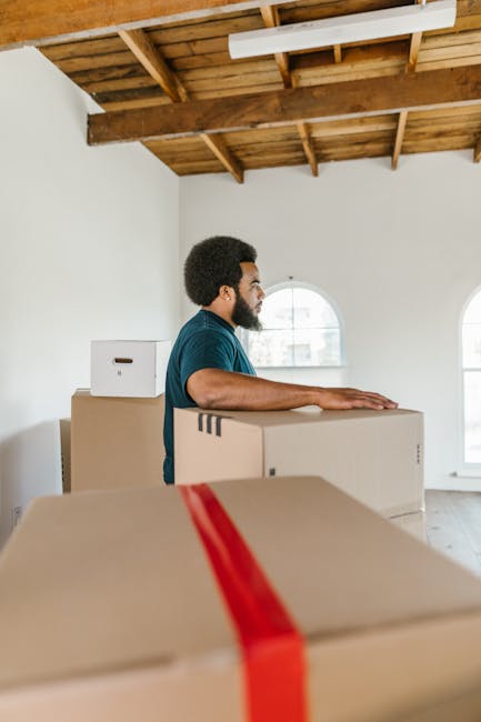 A man with dark curly hair and a beard, wearing a teal shirt, is shown in a spacious, well-lit interior with a wooden ceiling and large arched windows, participating in a home relocation process. He is standing behind a stack of cardboard moving boxes, which are closed and secured with packing tape, some of which are placed on the floor while others are stacked at waist height. One of the boxes in the foreground has a red strap around it, possibly for secure transport. The move appears to be in progress within a residential space, likely preparing for furniture transport or packing and moving activities. Removal Companies Harringay, operating in the area, specializes in house removals, including packing, loading, and logistics, as illustrated by the ongoing process of handling boxes for a house move or student relocation in Harringay N4. The environment suggests a professional approach to home relocation, with a focus on storing and organizing household items before transportation.