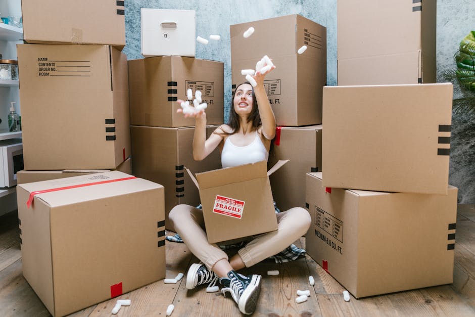 A young woman sitting cross-legged on a wooden floor inside a room, surrounded by numerous large cardboard moving boxes in various sizes, some sealed with red tape and labeled with handling instructions. She is smiling and tossing white packing peanuts into the air from a partially opened box in her lap. The background features a blue textured wall, a small white cabinet with a potted plant on top, and a green plant in the corner. The scene captures the process of home relocation, with a focus on packing and preparing items for a move, which is consistent with furniture transport and loading activities undertaken by Removal Companies Harringay as part of their house removals service.