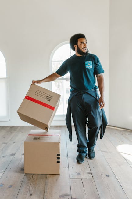 A man with dark curly hair and a beard, wearing a teal shirt, is shown in a spacious, well-lit interior with a wooden ceiling and large arched windows, participating in a home relocation process. He is standing behind a stack of cardboard moving boxes, which are closed and secured with packing tape, some of which are placed on the floor while others are stacked at waist height. One of the boxes in the foreground has a red strap around it, possibly for secure transport. The move appears to be in progress within a residential space, likely preparing for furniture transport or packing and moving activities. Removal Companies Harringay, operating in the area, specializes in house removals, including packing, loading, and logistics, as illustrated by the ongoing process of handling boxes for a house move or student relocation in Harringay N4. The environment suggests a professional approach to home relocation, with a focus on storing and organizing household items before transportation.