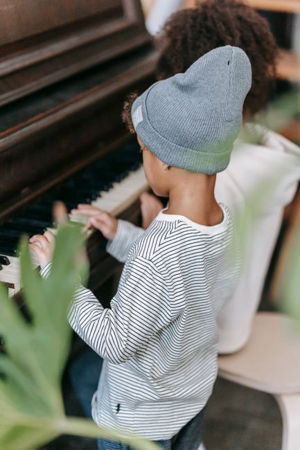 A young child wearing a grey knit beanie and a white and black striped long-sleeve shirt is standing beside an adult, both watching as the child’s hand gently presses the keys of a vintage upright piano with a dark wooden finish. The piano is positioned indoors, near a window, with visible piano keys and part of the sheet music stand. The scene depicts a home environment, with the focused action of playing or exploring the piano, which may be part of a house relocation or packing process handled by Removal Companies Harringay. Surrounding elements include a glimpse of a green plant in the foreground and soft lighting that highlights the furniture and the activity inside the room.