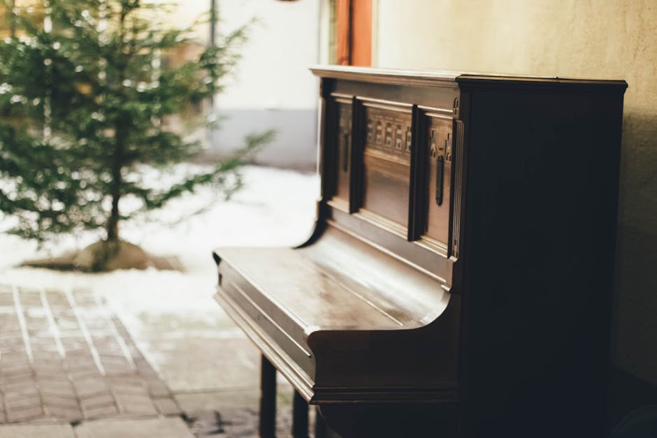 A vintage wooden upright piano with a polished dark finish is positioned indoors near a window, with natural light illuminating its surface. The piano's lid is closed, revealing detailed carvings and metal handles on its front panel. It is situated on a tiled floor, close to a doorway leading outside. Nearby, a small potted plant with green foliage is visible, adding a touch of nature to the scene. In the background, a beige wall and curtains can be seen, indicating a residential setting. The surrounding environment suggests a home undergoing packing or furniture transport, with the piano prepared for a professional removal by [COMPANY_NAME]. This scene reflects typical home relocation or furniture moving processes, emphasizing careful handling and spatial arrangements involved in house removals and furniture transport.