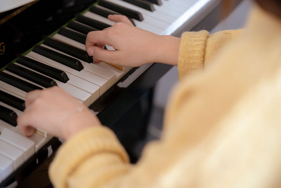 A young child wearing a grey knit beanie and a white and black striped long-sleeve shirt is standing beside an adult, both watching as the child’s hand gently presses the keys of a vintage upright piano with a dark wooden finish. The piano is positioned indoors, near a window, with visible piano keys and part of the sheet music stand. The scene depicts a home environment, with the focused action of playing or exploring the piano, which may be part of a house relocation or packing process handled by Removal Companies Harringay. Surrounding elements include a glimpse of a green plant in the foreground and soft lighting that highlights the furniture and the activity inside the room.